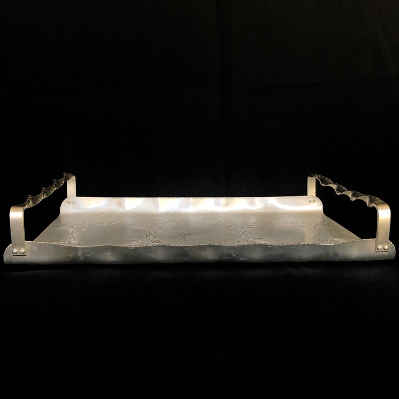 May include: A silver serving tray with decorative embossed details and two handles on each side. The tray is made of aluminum and has a slightly wavy edge.