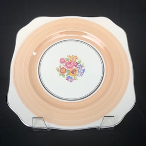 May include: A square, white ceramic plate with a peach-colored rim and a silver rim around the center. The center features a floral design with pink, yellow, blue, and green flowers.