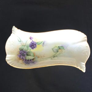 Pode incluir: Uma travessa retangular de porcelana branca com um design ondulado e acabamento dourado. A travessa é pintada à mão com violetas roxas e folhas verdes. O fundo tem um efeito suave de aquarela amarela e bege.