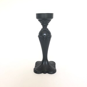 May include: A black candle holder with an ornate design. The holder has a wide base and a tall, slender stem.