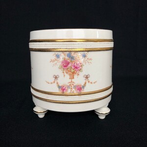 Op de afbeelding: Witte keramische plantenpot met een bloemenmotief en gouden rand. De pot heeft drie poten en is perfect voor het tentoonstellen van planten of bloemen.