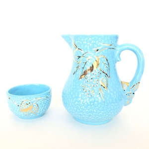 May include: A light blue ceramic pitcher and bowl set with gold leaf floral designs. The pitcher has a wide mouth and a handle. The bowl is round and has a small lip.