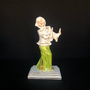 May include: A ceramic figurine of a woman wearing a white and gold patterned dress and a green skirt. She is standing on a white pedestal with gold trim.
