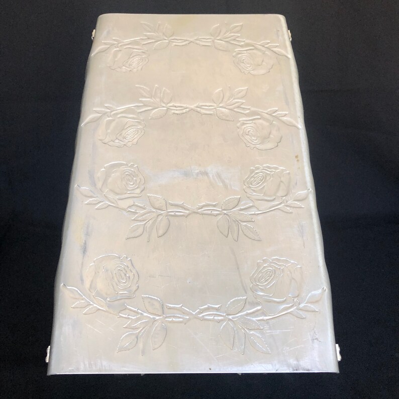 May include: A silver metal box with a decorative design of roses and vines. The box has a rectangular shape and a slightly curved top.