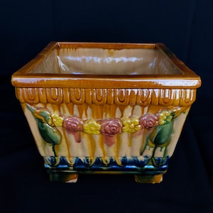 May include: A square ceramic planter with a floral design. The planter is decorated with yellow, pink, and green flowers and leaves. The planter has a brown glaze and a blue and white trim.