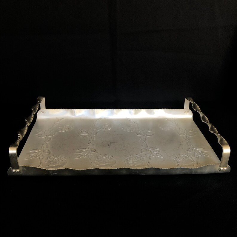 May include: A silver serving tray with a wavy edge and embossed rose design. The tray has two handles on either side.