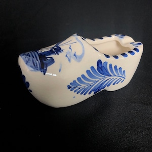 Może przedstawiać: Biały ceramiczny chodak holenderski, zdobiony niebieskimi motywami kwiatowymi i wiatrakami. Ten dekoracyjny but, o zakrzywionym kształcie, jest prawdopodobnie pamiątką lub elementem wystroju domu.