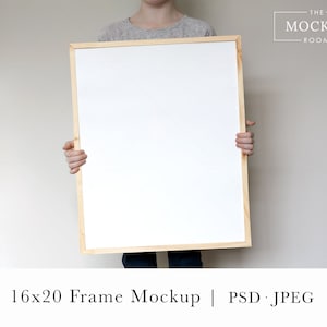 May include: A person holds a 16x20 inch wood frame mockup with a white background. The frame is made of natural wood and has a simple design.