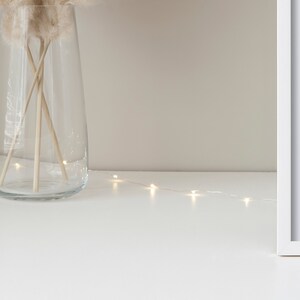 May include: A clear glass vase with dried pampas grass and a string of white fairy lights on a white surface.