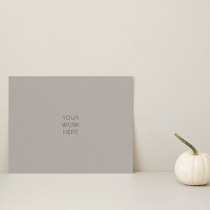 May include: A gray rectangular sign with the text "YOUR WORK HERE" is propped up on a white surface. A small white pumpkin with a green stem sits to the right of the sign. The background is a neutral beige.