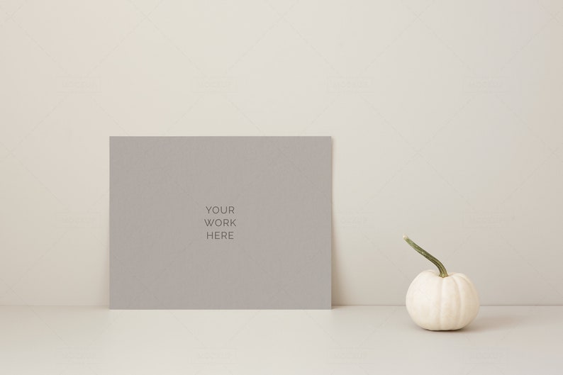 May include: A gray rectangular sign with the text "YOUR WORK HERE" is propped up on a white surface. A small white pumpkin with a green stem sits to the right of the sign. The background is a neutral beige.