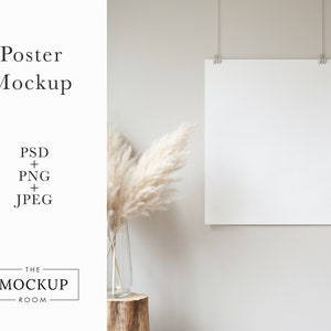 May include: A white poster mockup hanging on a light beige wall with a vase of dried pampas grass in front of it. The poster is hanging from two strings with clips.