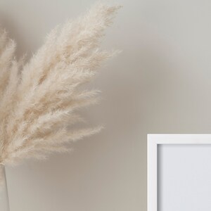 May include: A white picture frame with a white mat is on a light beige wall. A vase with dried pampas grass is in the foreground.
