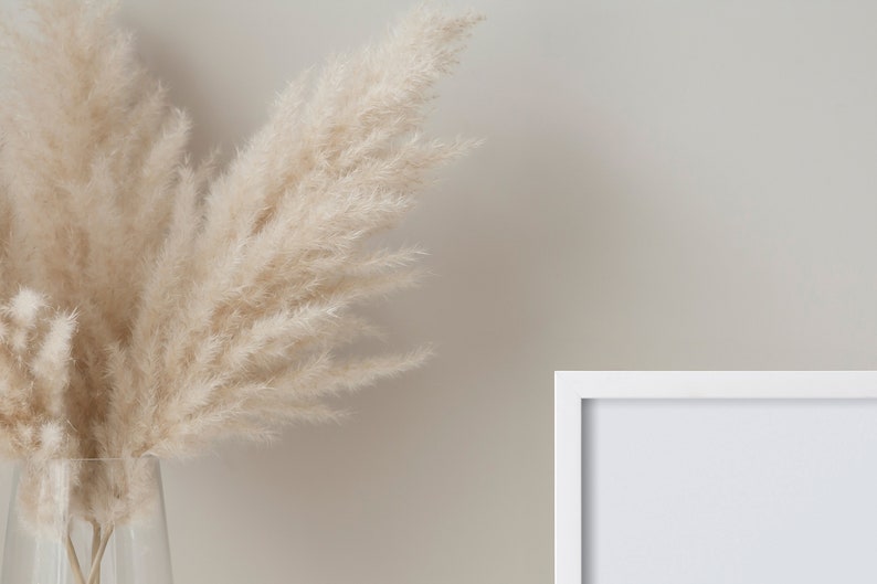 May include: A white picture frame with a white mat is on a light beige wall. A vase with dried pampas grass is in the foreground.