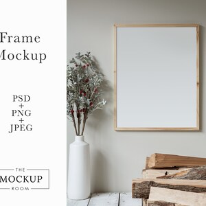 May include: A wooden frame mockup with a white background, a white vase with a sprig of greenery and red berries, and a pile of wood logs in the foreground.