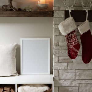 May include: A white picture frame with a blank canvas sits on a white shelf with a white faux fur throw in a wicker basket below. A wooden shelf above features a gold deer figurine and a glass candle holder with a lit candle. Two red and white Christmas stockings hang from a stone fireplace.