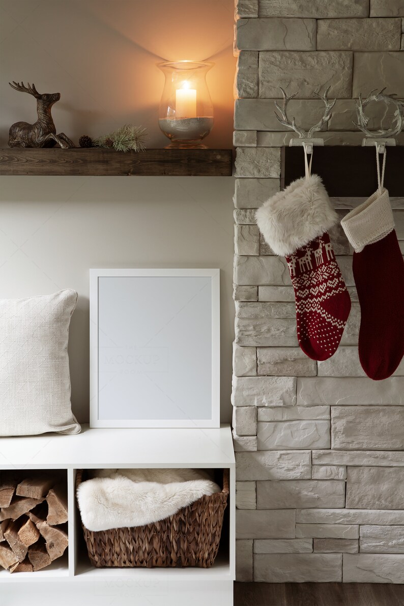 May include: A white picture frame with a blank canvas sits on a white shelf with a white faux fur throw in a wicker basket below. A wooden shelf above features a gold deer figurine and a glass candle holder with a lit candle. Two red and white Christmas stockings hang from a stone fireplace.