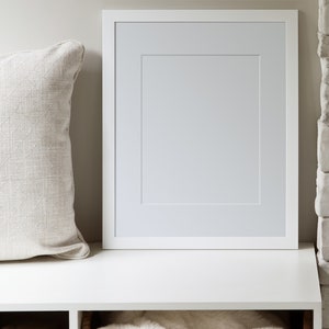 May include: A white picture frame with a white mount is on a white shelf. A white throw blanket is in a wicker basket below the shelf. A stack of firewood is in the shelf to the left of the basket.
