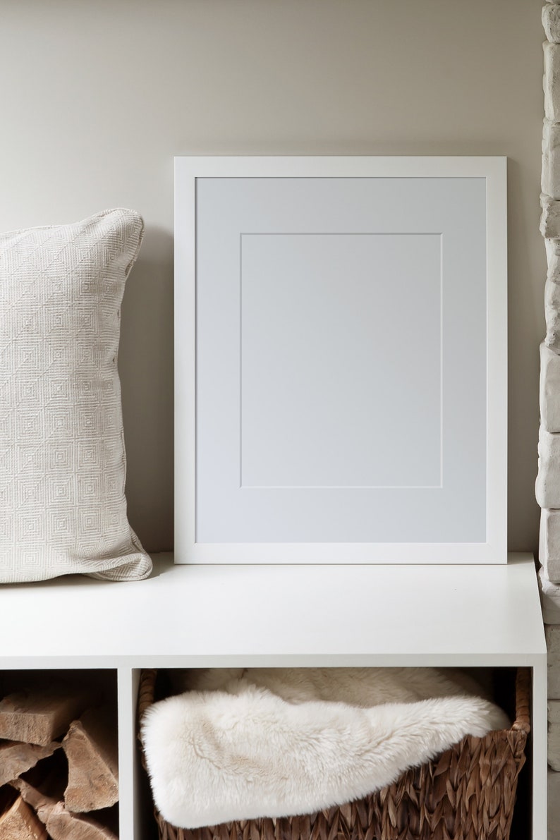 May include: A white picture frame with a white mount is on a white shelf. A white throw blanket is in a wicker basket below the shelf. A stack of firewood is in the shelf to the left of the basket.