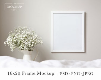 May include: A white 16x20 inch frame mockup with a white mat, hanging on a white wall. A white pitcher with a bouquet of white baby's breath flowers sits on a white bedspread.