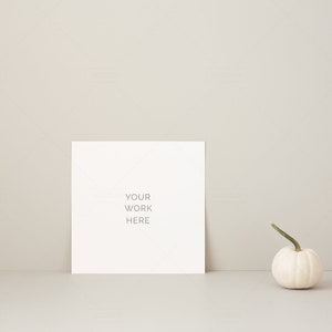 May include: A white square card with the text "YOUR WORK HERE" stands upright. A small white pumpkin with a green stem is to the right. The background is a neutral beige color.
