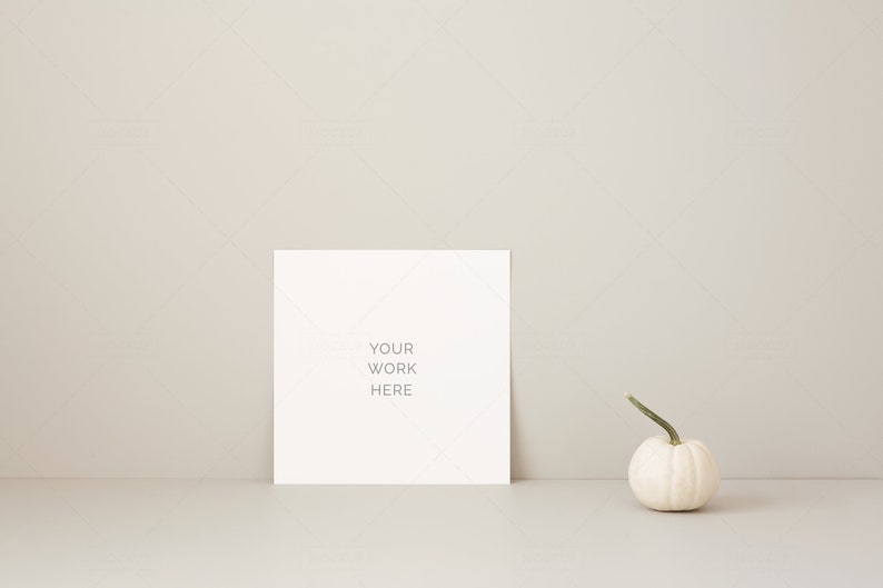 May include: A white square card with the text "YOUR WORK HERE" stands upright. A small white pumpkin with a green stem is to the right. The background is a neutral beige color.