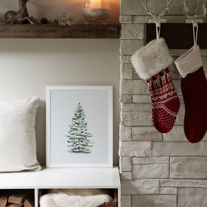 May include: A white shelf with a framed watercolour painting of a green pine tree. The shelf is above a white storage bench with a wicker basket filled with a white faux fur throw. The bench is in front of a stone fireplace with two red and white knitted stockings hanging from hooks.