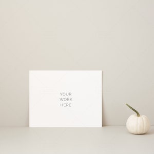 May include: A white card with the text "YOUR WORK HERE" is propped up against a neutral-colored wall. A small white pumpkin with a green stem sits to the right of the card. The scene is minimalist and evokes a fall theme.