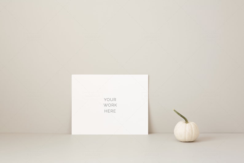 May include: A white card with the text "YOUR WORK HERE" is propped up against a neutral-colored wall. A small white pumpkin with a green stem sits to the right of the card. The scene is minimalist and evokes a fall theme.