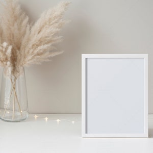 May include: A white picture frame with a blank white background sits on a white surface with a vase of dried pampas grass and a string of white fairy lights.