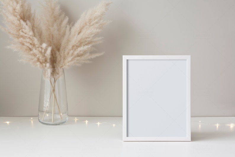 May include: A white picture frame with a blank white background sits on a white surface with a vase of dried pampas grass and a string of white fairy lights.