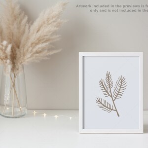 May include: A white frame with a brown line drawing of a pine branch on a white background. The frame is sitting on a white surface with a vase of dried pampas grass in the background.