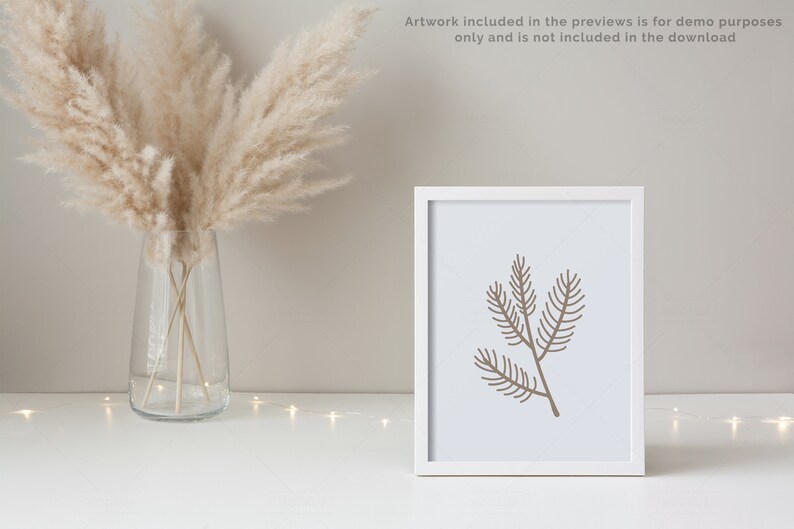 May include: A white frame with a brown line drawing of a pine branch on a white background. The frame is sitting on a white surface with a vase of dried pampas grass in the background.