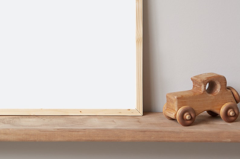May include: A wooden toy car sits on a wooden shelf in front of a blank picture frame. The car is made of natural wood and has a simple design.