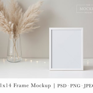 May include: White frame mockup with a white mat, sitting on a white surface with a vase of dried pampas grass and a string of fairy lights. The frame is 11x14 inches.