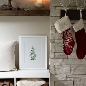 May include: A white shelf with a wooden top, a framed print of a green Christmas tree, a white faux fur throw in a wicker basket, and a stack of firewood in a cubby below. Two red and white Christmas stockings hang on a brick fireplace.