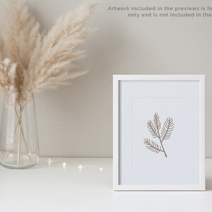 May include: A white frame with a simple line drawing of a pine branch inside. The frame is sitting on a white surface with a vase of dried pampas grass in the background.