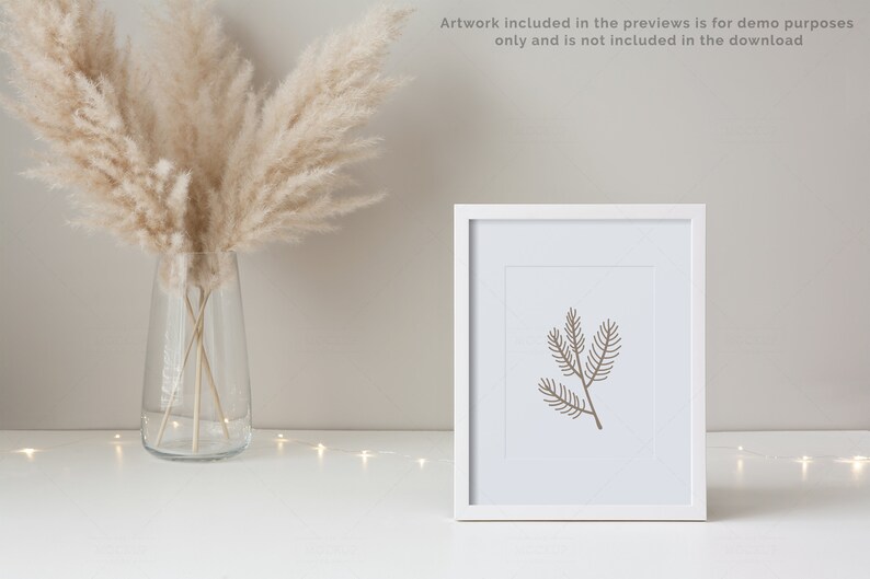 May include: A white frame with a simple line drawing of a pine branch inside. The frame is sitting on a white surface with a vase of dried pampas grass in the background.