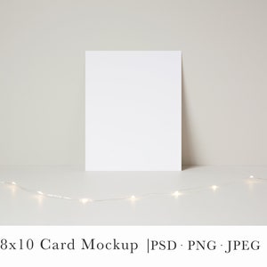 May include: A white 8x10 card mockup on a white surface with a string of white fairy lights in front of it.