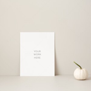 May include: A blank white rectangular sign with the text "YOUR WORK HERE" is propped up against a neutral-colored wall. A small white pumpkin with a green stem sits to the right of the sign. The scene is minimalist and autumnal.