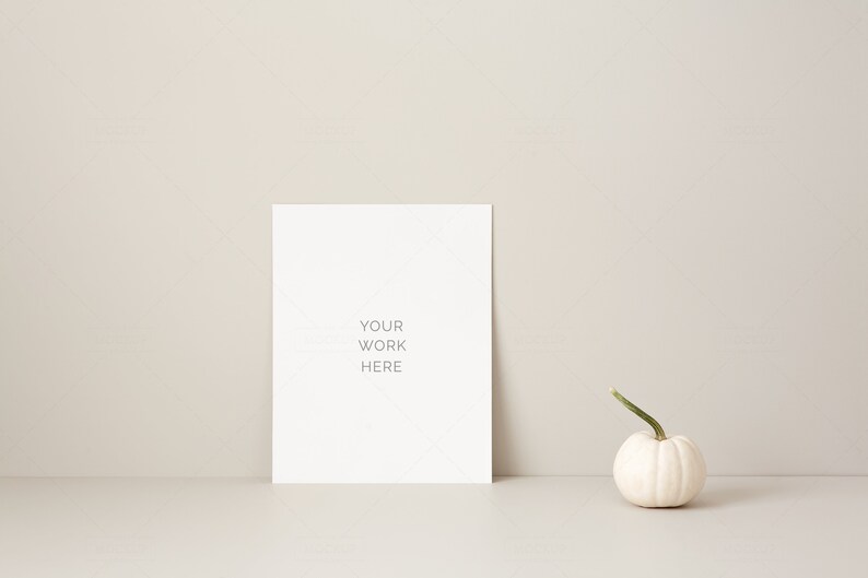 May include: A blank white rectangular sign with the text "YOUR WORK HERE" is propped up against a neutral-colored wall. A small white pumpkin with a green stem sits to the right of the sign. The scene is minimalist and autumnal.