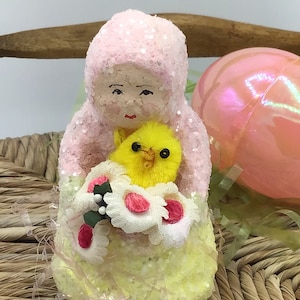 May include: A pink and yellow vintage Easter figurine of a girl holding a yellow chick and flowers. The figurine is made of cotton and covered in glitter.