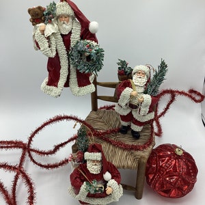 May include: Three red and white Santa Claus ornaments with white fur trim. One Santa is holding a teddy bear and a wreath, another is holding a small tree and a bag, and the third is holding a small tree and a bag of candy canes. The ornaments are sitting on a wooden chair with red tinsel.