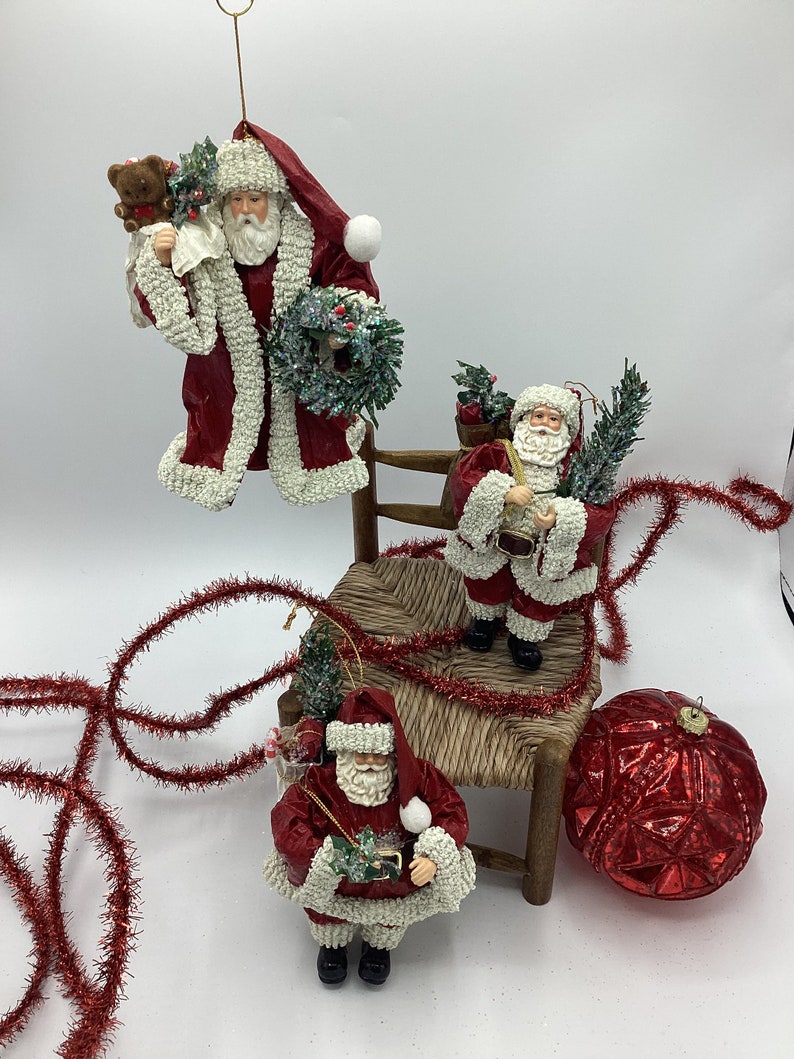 May include: Three red and white Santa Claus ornaments with white fur trim. One Santa is holding a teddy bear and a wreath, another is holding a small tree and a bag, and the third is holding a small tree and a bag of candy canes. The ornaments are sitting on a wooden chair with red tinsel.