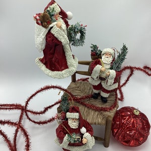 May include: Three red and white Santa Claus ornaments with white beards and hats. The Santas are holding a small green tree, a wreath, and a teddy bear. The ornaments are sitting on a small wooden chair with a woven seat. A red and white Christmas garland is in the background.