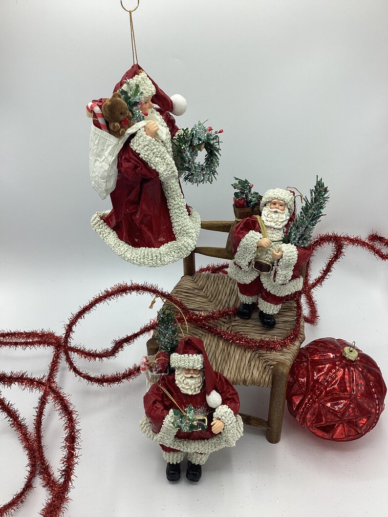 May include: Three red and white Santa Claus ornaments with white beards and hats. The Santas are holding a small green tree, a wreath, and a teddy bear. The ornaments are sitting on a small wooden chair with a woven seat. A red and white Christmas garland is in the background.
