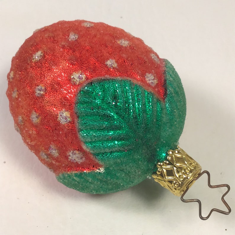 May include: A red and green glass Christmas bauble shaped like a strawberry with a gold star topper.