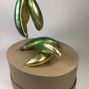 May include: Banana-shaped Christmas tree decorations in green and gold. The ornaments have a shiny, reflective finish. Some are suspended, others rest on a brown, circular base.