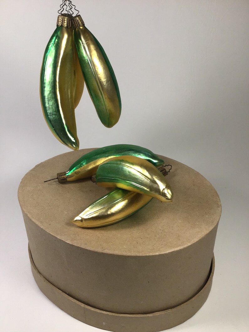 May include: Banana-shaped Christmas tree decorations in green and gold. The ornaments have a shiny, reflective finish. Some are suspended, others rest on a brown, circular base.