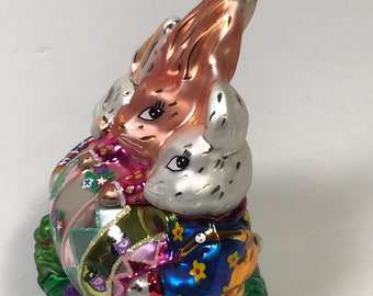 以下が含まれることがあります： 花の上に座っている2羽のウサギが描かれたガラス製のクリスマスオーナメント。ウサギはカラフルな服を着て、大きなカラフルな卵を持っています。オーナメントは、吊るすための金色のトップが付いています。
